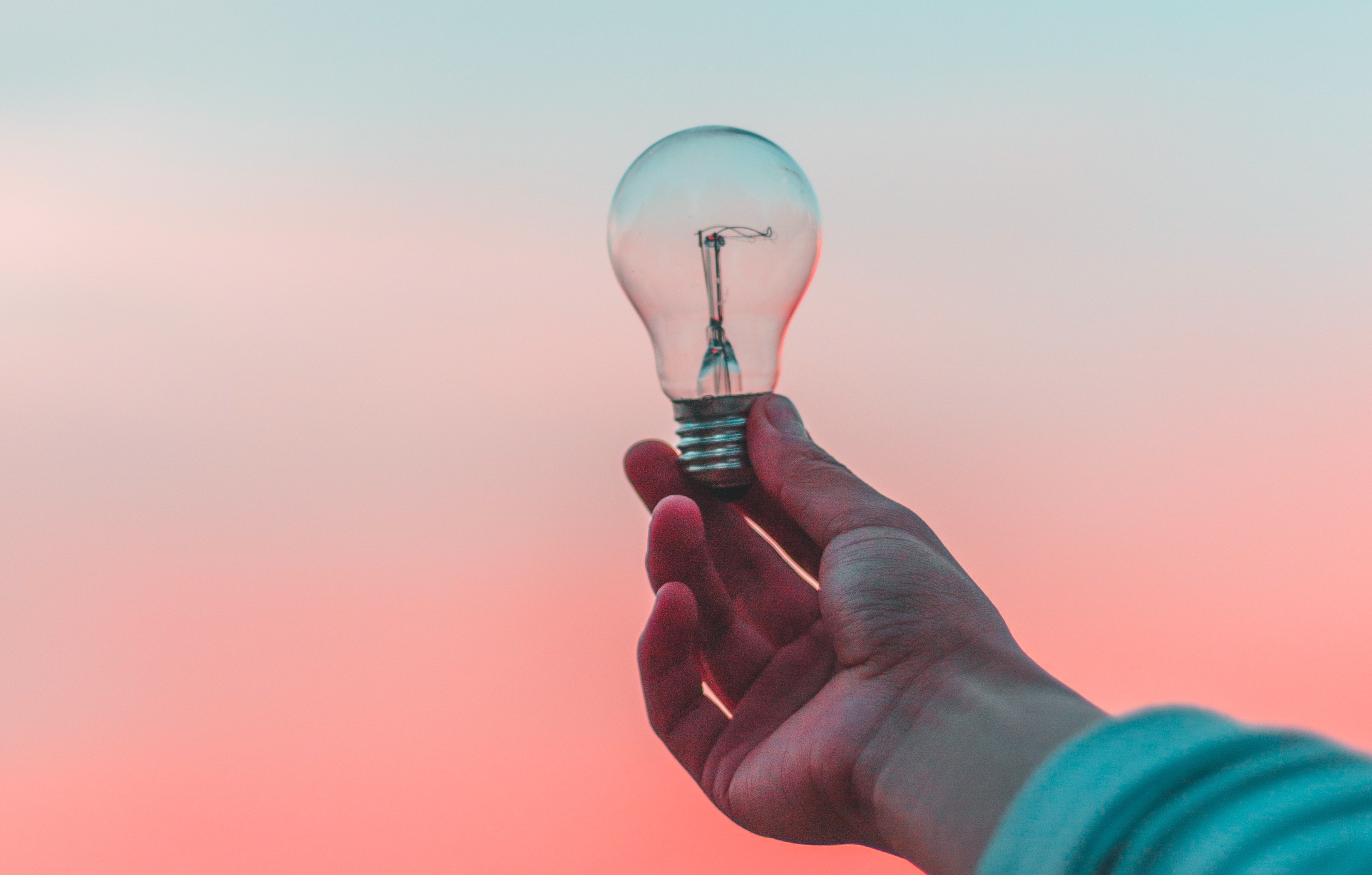 En hand håller upp en glödlampa mot en rosa himmel